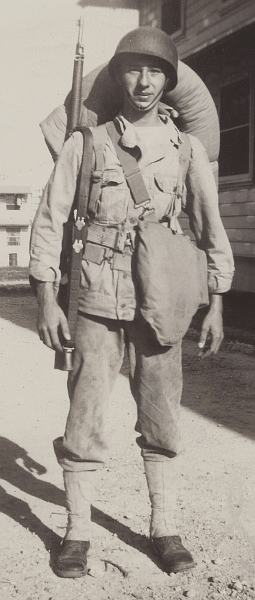 This 1942 photo shows Private William Zemba fully equipped for field training exercises.  He wears the two‐piece herringbone twill suit with the jacket bottom exposed, canvas leggings, gas mask carrier strapped to body, Springfield rifle on right shoulder, pistol belt with suspenders, full pack, and M‐1 helmet liner.