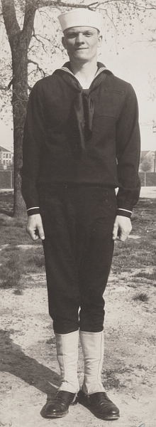 This 1942 photo shows a young Apprentice Seaman wearing the Dress Blue B uniform at his training station.  His jumper has one cuff stripe and is being worn bloused over at the waist in the prescribed manner.  His neckerchief is rolled and knotted at the base of the "V" on the jumper.  He wears high-top shoes and leggings.