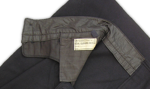 A view of the right, inside waistband of the 1944 trousers showing the loop of tape used to tie the trousers to a laundry line.  It is visible just above the Naval Clothing Factory label.  There were two tape loops, one located on each side of the trousers, which replaced the eyelets used on the old design.  The design of the new trousers was intended to simplify manufacture and reduce costs.
