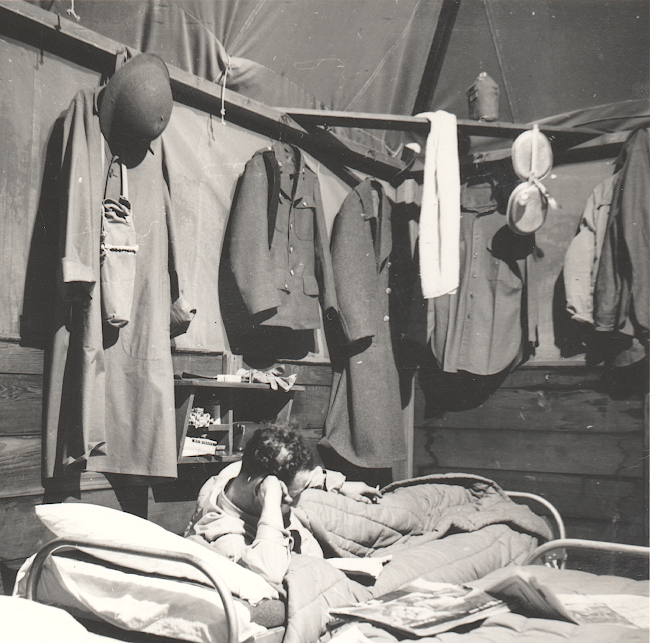 This photo shows a GI relaxing in his bunk with his wardrobe hanging overhead. From left to right are the M1917‐A1 steel helmet, gas mask carrier, raincoat, wool service coat with leather belt draped over the right shoulder, wool overcoat, flannel shirt with front button placket, and partially visible at far right is the olive‐drab field jacket and another flannel shirt. On the corner cross beam is a towel, canteen, and mess kit. On the top shelf above the man's head lay his garrison cap and a pair of work gloves.
